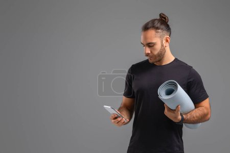 A man standing and holding a yoga mat in one hand and a cell phone in the other, ready for a workout or yoga session, possibly checking his phone for instructions or tracking his progress, copy space
