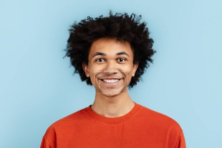 Porträt eines fröhlichen afrikanisch-amerikanischen Typen mit einem einnehmenden Lächeln, der einen orangefarbenen Pullover trägt und vor blauem Hintergrund posiert. Stellt Glück und Jugendlichkeit dar