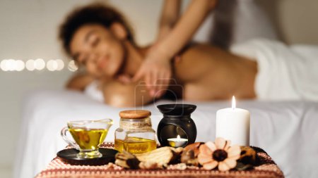 Photo for In-focus spa oil and blurred background of an african american woman receiving a massage at a spa with a happy expression - Royalty Free Image