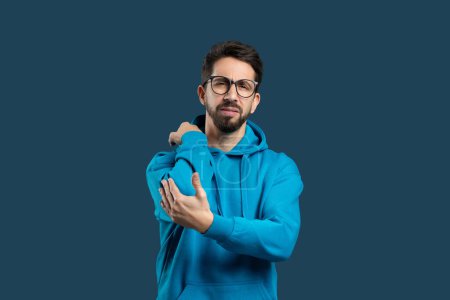 A young man wearing glasses and blue hoodie stands against plain blue backdrop, touching elbow with pained expression that suggests he may be experiencing discomfort or injury in that area.