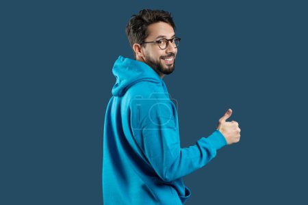 A man wearing a blue hoodie is shown in the image, raising his hand to give a thumbs up gesture. The mans face is not visible, and he appears to be standing against a plain background.