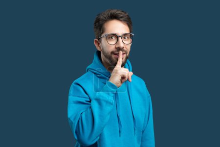 Photo for A man in a bright blue hoodie stands against a matching blue background, placing one finger over his lips as if to gesture for silence or secrecy. - Royalty Free Image