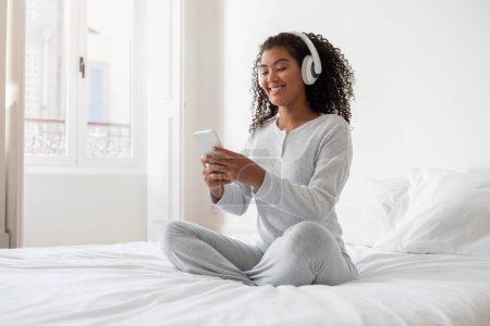 Photo for A young Hispanic woman sits cross-legged on a pristine white bed in a sunlit bedroom, smiling as she listens to music through her white headphones. She holds and attentively looks at her smartphone - Royalty Free Image