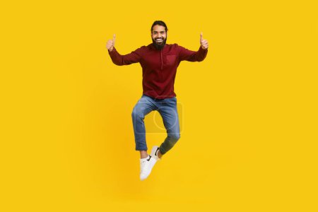 Indian man is captured mid-air, jumping energetically with his hands raised in thumb ups gesture, yellow studio background
