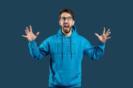 Photo for A delighted young man with glasses is standing against a dark blue backdrop, his mouth wide open as if shouting with joy or surprise. He is wearing a bright blue hoodie and has raised his hands - Royalty Free Image