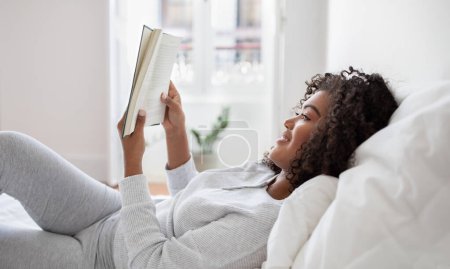 A Hispanic woman is lying down in bed with a book in hand, engrossed in reading. The room is dimly lit, and she appears relaxed and focused on the pages of the book, side view