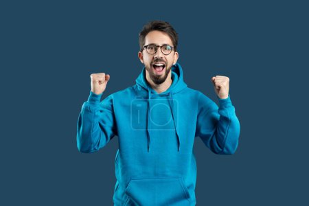 Un homme en sweat à capuche bleu avec ses poings levés célébrant le succès, montrant des émotions positives isolées sur fond de studio bleu