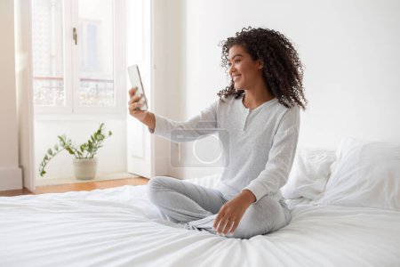 Foto de Una mujer hispana está sentada en una cama, sosteniendo su teléfono para tomar una selfie. Ella está sonriendo y mirando la pantalla del teléfono mientras captura el momento. - Imagen libre de derechos