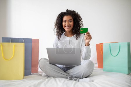 Une jeune femme hispanique joyeuse s'assoit sur son lit avec un ordinateur portable, affichant une carte de crédit avec des sacs à provisions autour d'elle, suggérant une séance de magasinage en ligne réussie.