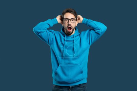 A young man wearing blue hoodie and glasses stands with his mouth open in shock, hands placed on his head against plain blue background, expressing surprise or disbelief at unexpected situation.