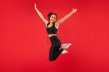 Photo for Cheerful woman wearing a black sports bra top and black leggings is captured mid-jump. She showcases agility and strength as she leaps energetically. - Royalty Free Image