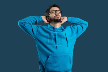 A young man wearing glasses and a casual blue hoodie stands with his hands behind his head, eyes closed in a gesture of relaxation and contentment, blue background