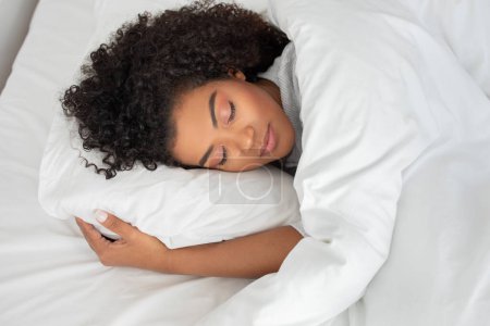 Eine hispanische Frau liegt mit geschlossenen Augen auf einem Bett und ruht friedlich. Ihr Körper ist entspannt und sie scheint sich in einem Zustand des Tiefschlafs oder der Entspannung zu befinden..