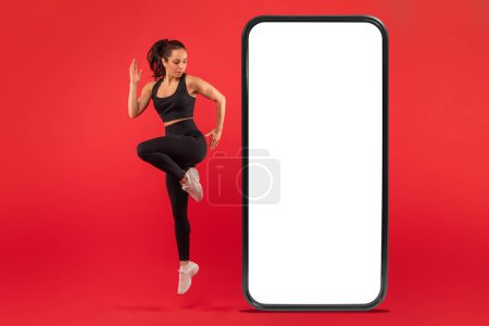 Eine in Sportkleidung gekleidete Frau wird in der Mitte der Bewegung eingefangen, während sie neben einem hoch aufragenden Smartphone mit leerem Bildschirm eine Übung mit hohen Knien ausführt.