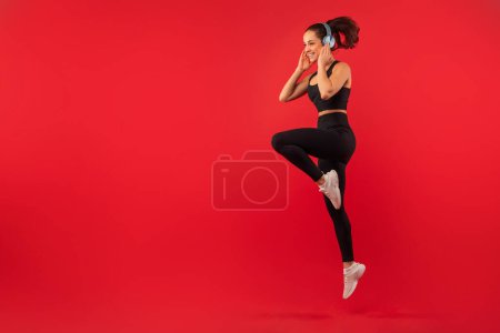 A joyful young woman with her hair tied in a ponytail is captured mid-jump against a bold red backdrop, showcasing her active lifestyle, using wireless headphones, copy space