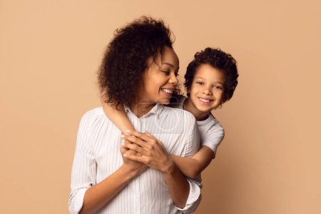 A smiling African American woman and her cheerful young son share a tender embrace, both sporting curly hair and wearing striped shirts, warm beige backdrop.