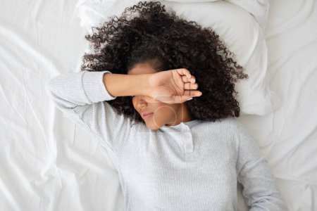 Une jeune femme est allongée sur un couvre-lit blanc, couvrant son visage de son bras à son réveil. La lumière naturelle du matin suggère le début d'une nouvelle journée