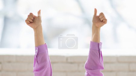 Foto de Dos brazos se levantan en aprobación, cada uno dando un gesto de pulgar hacia arriba. La mujer lleva una camisa morada de manga larga, y el fondo está suavemente fuera de foco. - Imagen libre de derechos