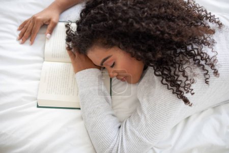 Téléchargez les photos : Une jeune femme hispanique aux cheveux bouclés repose sur un lit blanc, la tête posée sur un livre ouvert. Elle semble prendre une pause ou s'est peut-être endormie en lisant. - en image libre de droit