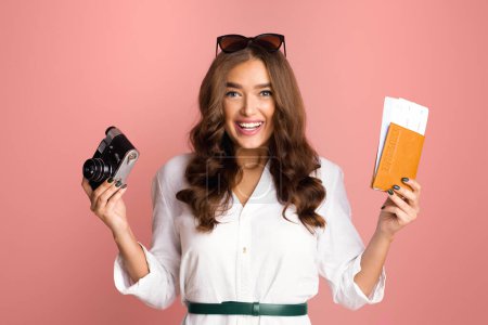 A cheerful young woman with wavy hair is holding a classic black camera in one hand and an orange airplane ticket or boarding pass in the other, against a solid pink backdrop