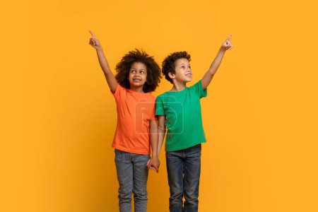 Photo for A pair of cheerful young African American friends stands side by side against a vibrant yellow backdrop, with both boys looking upwards and pointing excitedly at something above. - Royalty Free Image