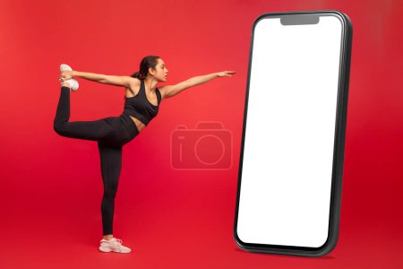 Flexible junge Frau in schwarzer Sportbekleidung, die neben einem riesigen Telefon mit weißem Bildschirm auf rotem Studiohintergrund übt, Yoga oder Pilates praktiziert, in voller Länge