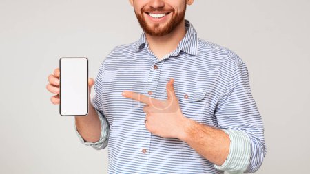 Photo for Smiling millennial man holding latest slim cellphone with blanc screen and pointing at it, panorama, free space - Royalty Free Image