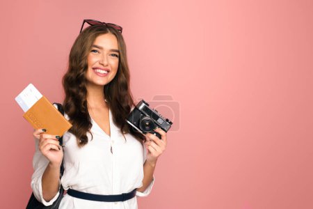 Eine gut gelaunte Frau mit Sonnenbrille auf dem Kopf zeigt Reisebereitschaft, während sie einen Pass und eine Oldtimer-Kamera in der Hand hält. Sie steht vor einem schlichten rosa Hintergrund