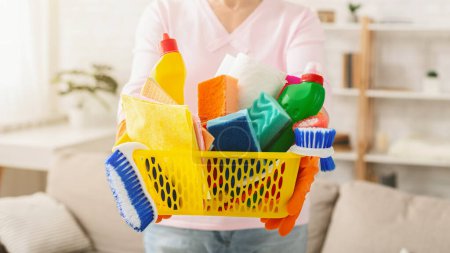 Cropped of woman stands in a sunlit living room, holding a yellow basket filled with various cleaning products and supplies. She is prepared for a housekeeping routine
