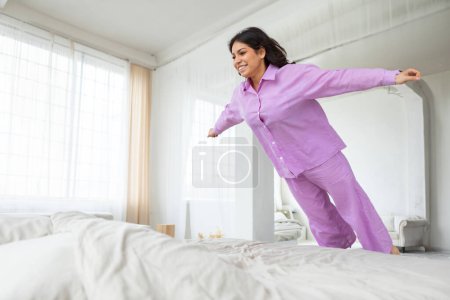 Photo for A middle eastern woman dressed in purple pajamas is energetically jumping on a white bed. The room is simple, with plain walls and a few scattered pillows - Royalty Free Image