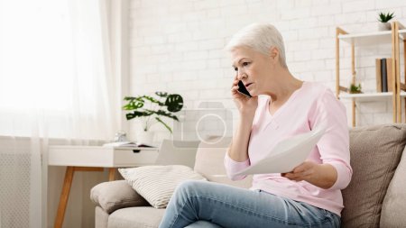 Wütende Seniorin telefoniert und arbeitet zu Hause, schaut auf Laptop und winkt mit Papier, sitzt zu Hause, freier Raum
