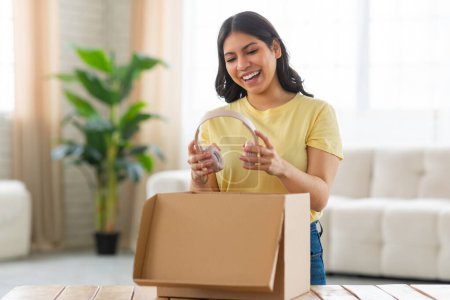 Photo for A young middle eastern woman stands in her sunlit living room, filled with excitement and a bright smile as she unpacks a cardboard box on a table, holding headphones - Royalty Free Image