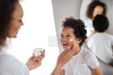 Ein kleines Kind, das vor Freude lacht, erhält sanfte Pflege von seiner Mutter, während sie Feuchtigkeitscreme auf seine Wange aufträgt. Afroamerikanische Familie steht dicht an dicht in einem gut beleuchteten Badezimmer