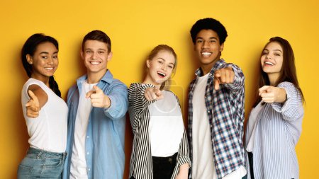 Photo for Multiethnic teenagers are standing together, smiling and pointing directly at the viewer. They are casually dressed and appear upbeat. The bright yellow background accentuates the joyful ambiance. - Royalty Free Image