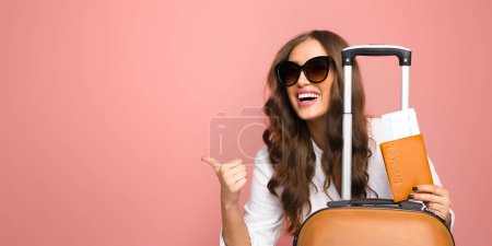 Foto de Una joven alegre con gafas de sol grandes está de pie con su equipaje, dando un cartel con los pulgares hacia arriba. Ella está vestida casualmente, lista para un viaje cómodo y elegante, espacio para copiar - Imagen libre de derechos