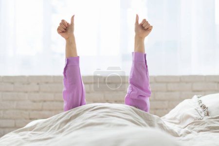 Téléchargez les photos : La femme est allongée sur un lit avec les bras tendus vers le haut, donnant un geste de pouce vers le haut avec les deux mains. La pièce est remplie de lumière naturelle, ce qui suggère qu'il fait jour. L'accent sur les mains - en image libre de droit