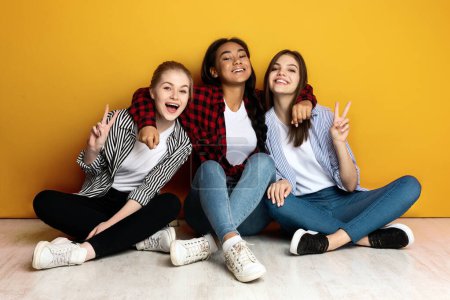 Drei junge multiethnische Mädchen sitzen dicht an dicht mit dem Rücken vor einer leuchtend gelben Wand. Sie sind lässig in Jeans und Flanellhemden gekleidet und lächeln fröhlich