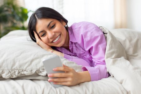 Téléchargez les photos : Une jeune femme joyeuse du Moyen-Orient se couche confortablement au lit, portant une chemise rose, alors qu'elle aime naviguer sur son smartphone dans le calme doucement éclairé de la matinée - en image libre de droit