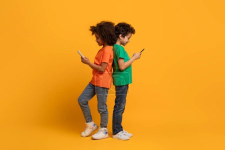 Photo for A pair of young African American children stand back-to-back, each absorbed in a smartphone. They are wearing casual clothing, against the vibrant yellow backdrop. - Royalty Free Image