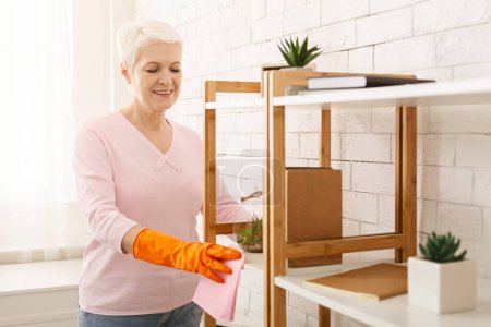 A cheerful elderly woman with short white hair is wearing orange gloves while wiping dust off her wooden shelves using a pink cloth. The sunlight illuminates the clean, white-walled room