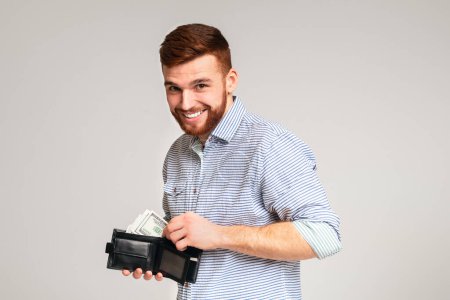 Finanzas, inversión y ahorro de dinero. Hombre guapo mostrando billetera negra con dólares y sonriendo, panorama, espacio libre
