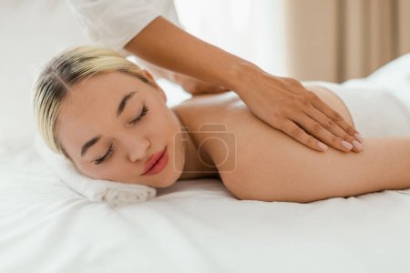 A woman lies face down on a massage table in a serene spa setting while a massage therapist performs a back massage on her. The therapists hands are visible as they apply pressure and knead