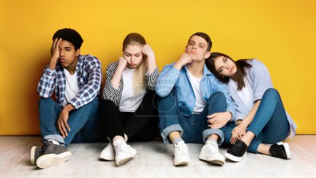 Vier multiethnische Teenager stehen vor einer leuchtend gelben Wand mit Ausdrücken von Langeweile und Erschöpfung. Sie wirken desinteressiert und unmotiviert, sitzen eng beieinander