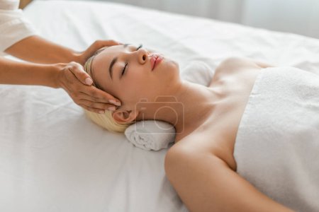Foto de Una mujer acostada con los ojos cerrados, recibiendo un suave masaje en la cara de un esteticista profesional con movimientos calmantes. - Imagen libre de derechos