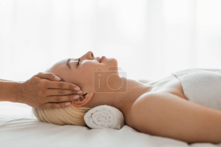 A woman lying down with her eyes closed as a professional masseuse massages her face, focusing on her forehead, cheeks, and jawline, side view