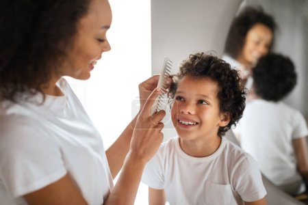 Photo for African American woman is seen combing a young boys hair in front of a mirror. Kid is sitting calmly as mother gently runs the comb through his hair, creating a bonding moment between them. - Royalty Free Image