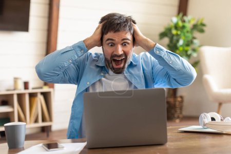 Ein Mann sitzt an einem Holztisch und drückt überwältigende Freude und Aufregung aus, während er auf seinen Laptop-Bildschirm blickt. Sein Mund steht staunend offen, Hände umklammern sein Haar.