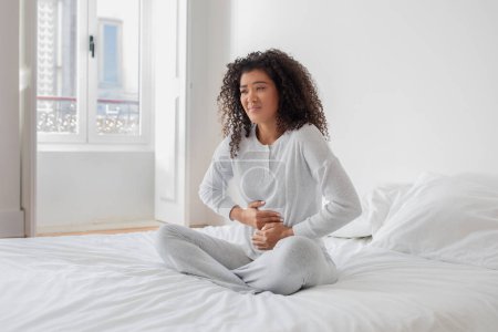 Mujer hispana joven usando pijamas sentada en la cama con las manos en el abdomen, sintiéndose mal por la mañana