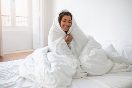 A smiling Hispanic woman is sitting on a bed, wrapped in a cozy blanket. She appears comfortable and content, exuding warmth and relaxation.