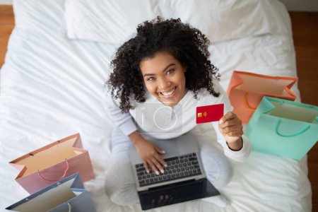 Foto de Una mujer está sentada en una cama, sosteniendo una tarjeta de crédito en una mano y una computadora portátil en la otra. Ella aparece enfocada y comprometida en una transacción en línea o actividad financiera, vista superior - Imagen libre de derechos